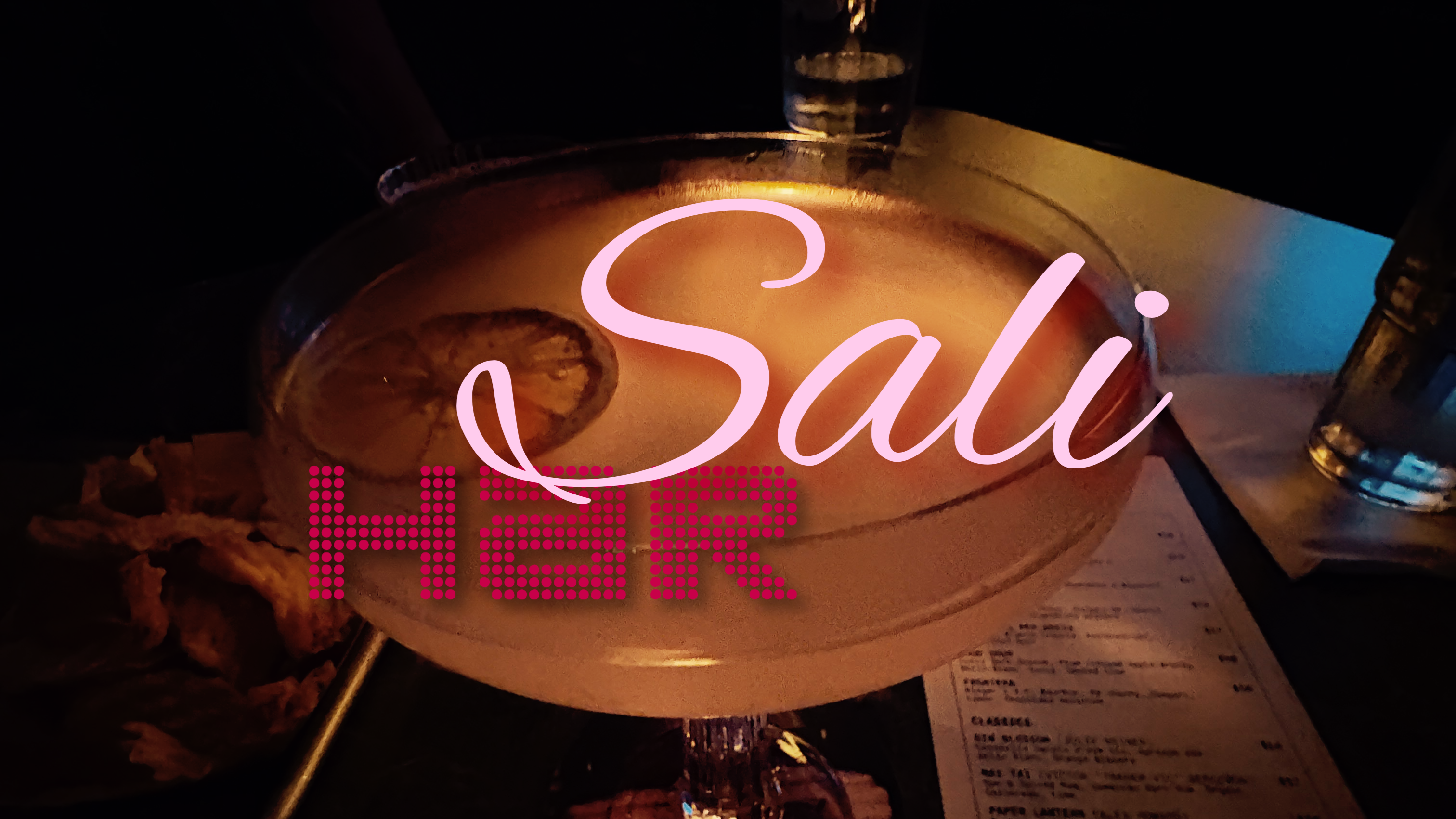A dimly lit cocktail in a large coupe glass on a table at a bar, with a neon sign reading 'harSali' overlaid across the front. A menu and other bar elements are visible in the dark surroundings.